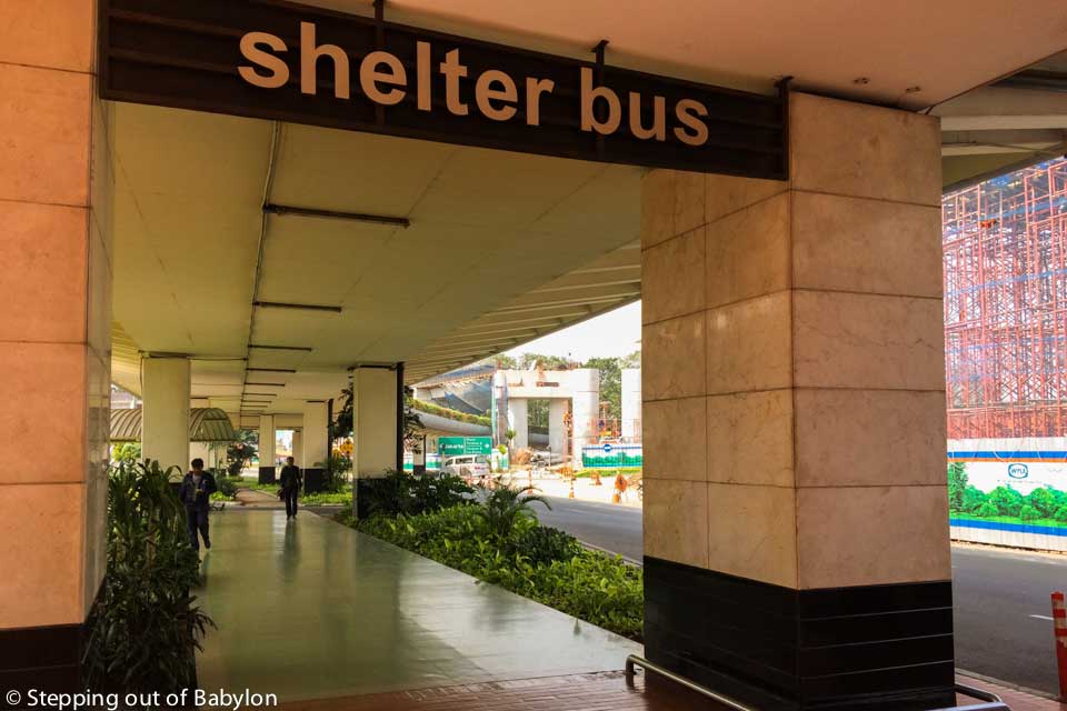 When you leave the airport building (arrivals) you must turn left, and keep walking under the roof until the end of the arrivals building. There you will see a sign (a kind of gate) with the words “Shelter Bus”.